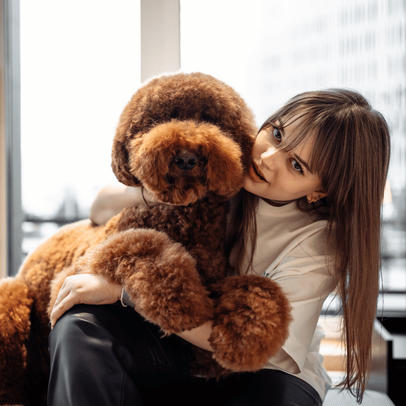 In the comfort of her home, a woman embraces her large, fluffy brown dog near a window, basking in shared affection.