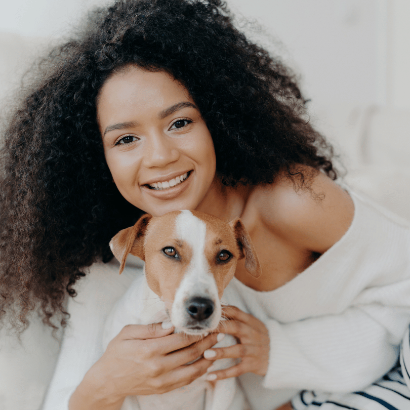 A woman with curly hair smiles while holding a small dog indoors.