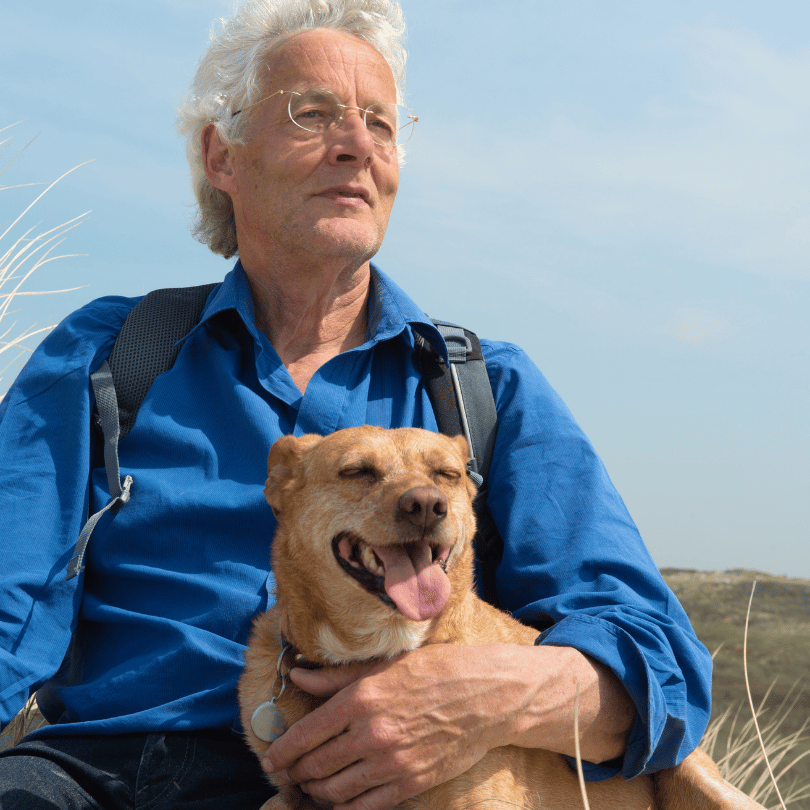 An older man in a blue shirt sits outside with a small brown dog on his lap. They're both looking into the distance on a sunny day.