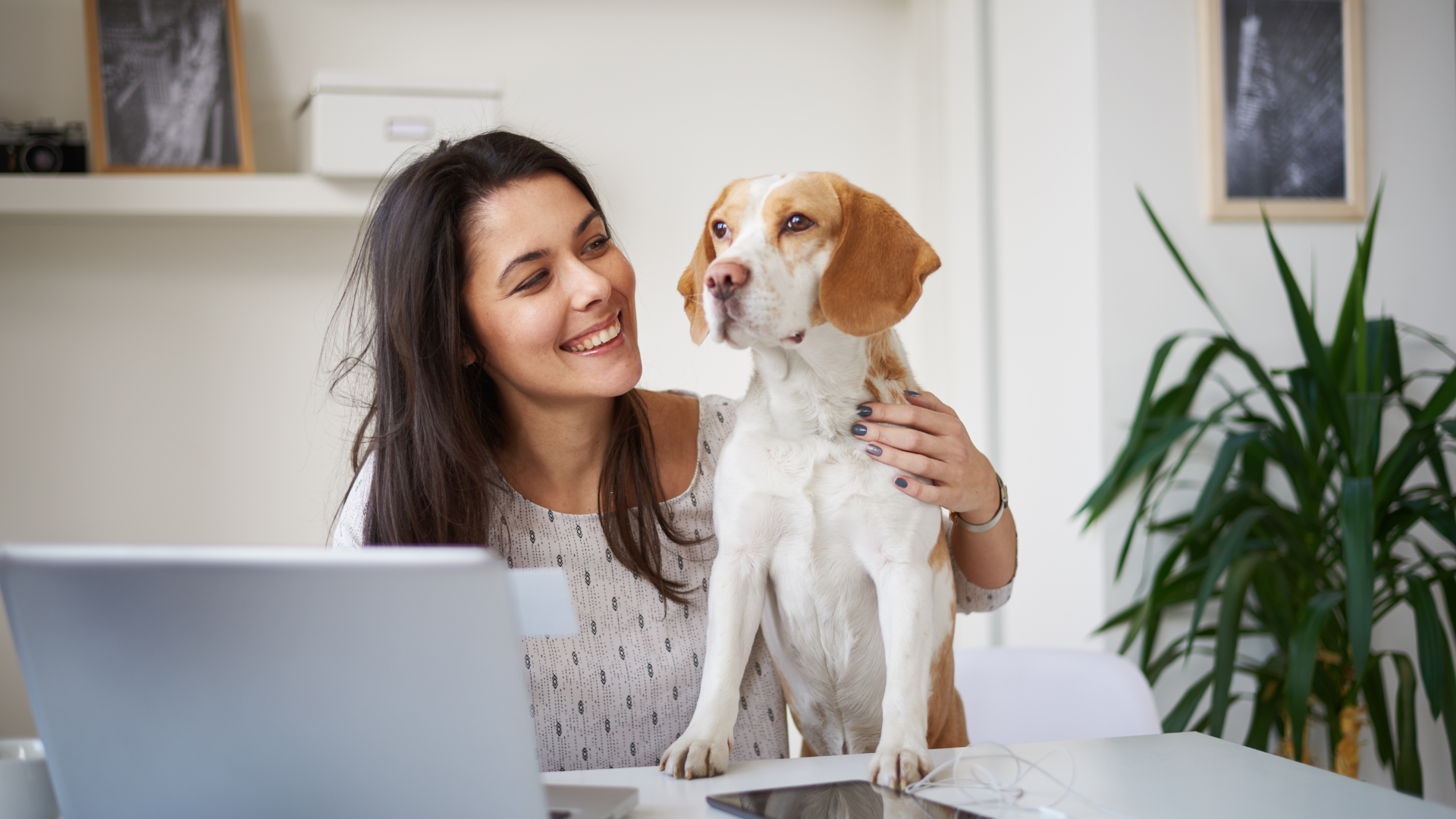 A woman smiling at a dog sitting beside her at a desk with a laptop, in a home office setting.