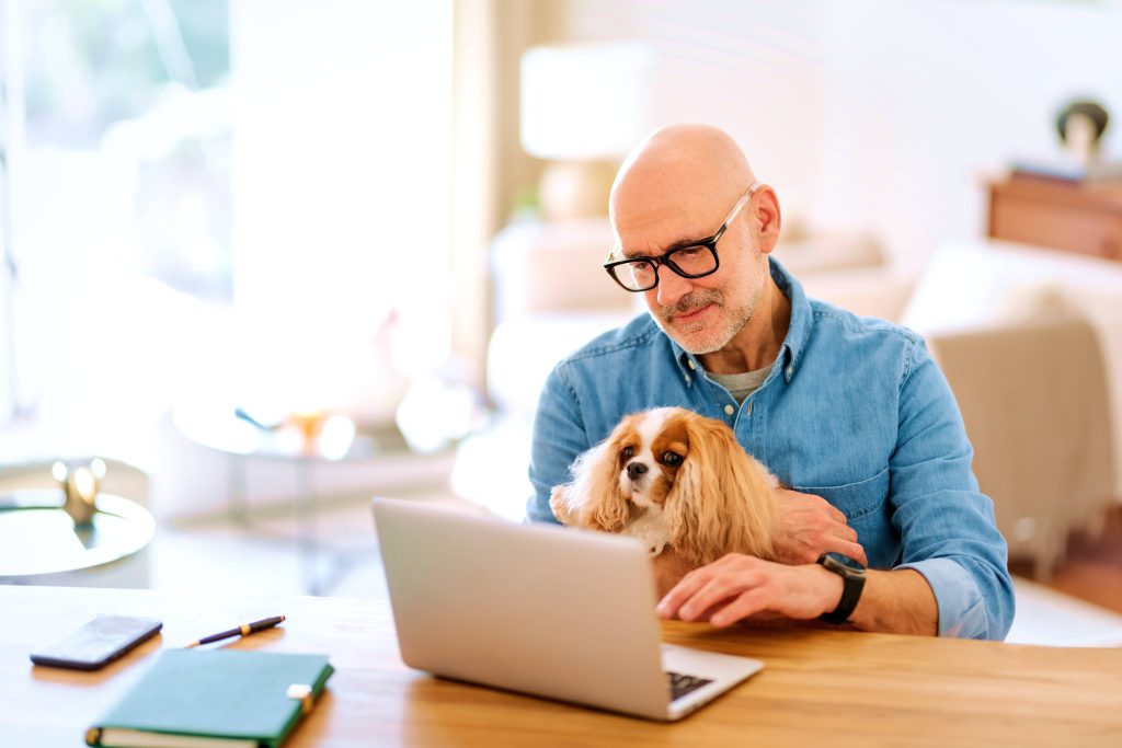 A man wearing glasses works on a laptop at a table, holding a small dog in his lap, showcasing his dedication to pet care.