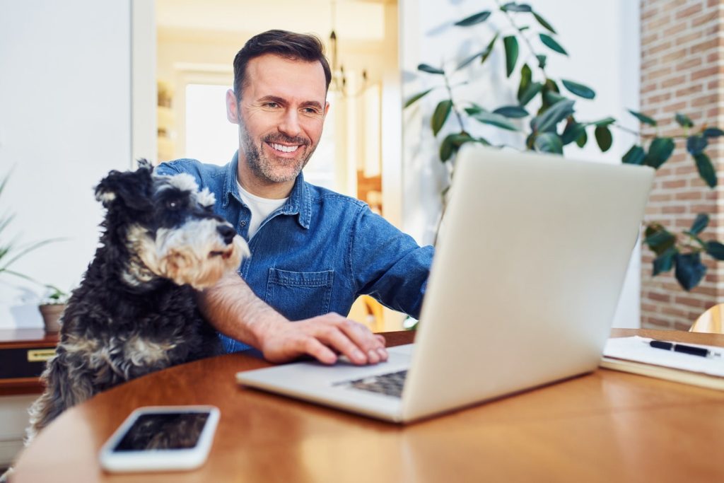 A man using a laptop at a wooden table with a smartphone nearby, while his black and white dog, fresh from grooming, sits beside him.