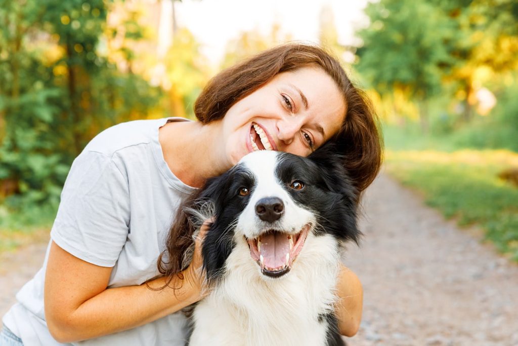 A woman with brown hair smiles as she hugs a black and white dog outside on a sunny day, with greenery in the background. Both appear happy, showcasing the bond that excellent pet care can foster.