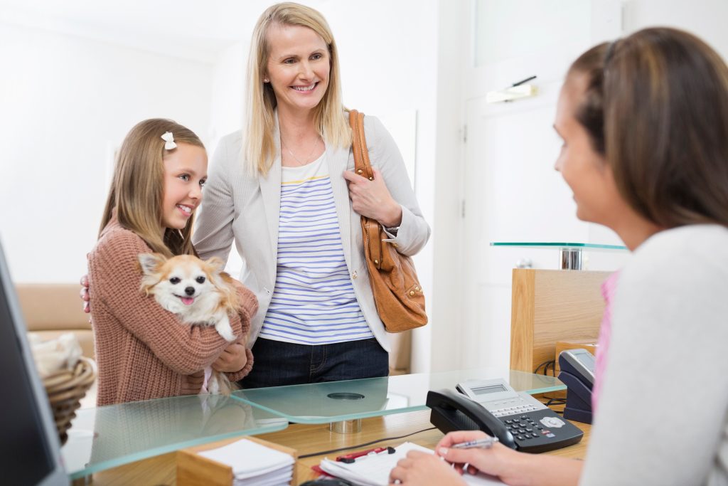 A woman and child holding a dog stand at a reception desk, smiling at the receptionist, ready to discuss pet care options.