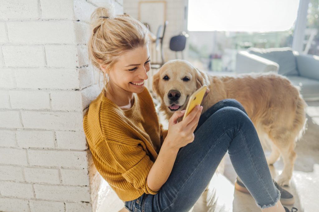 Woman smiling at smartphone with her golden retriever, using pet care app.