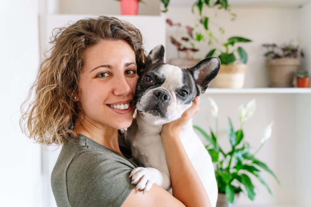 A woman with curly hair smiles while holding a black and white French Bulldog in a room with potted plants, showcasing their strong bond nurtured through attentive pet care.