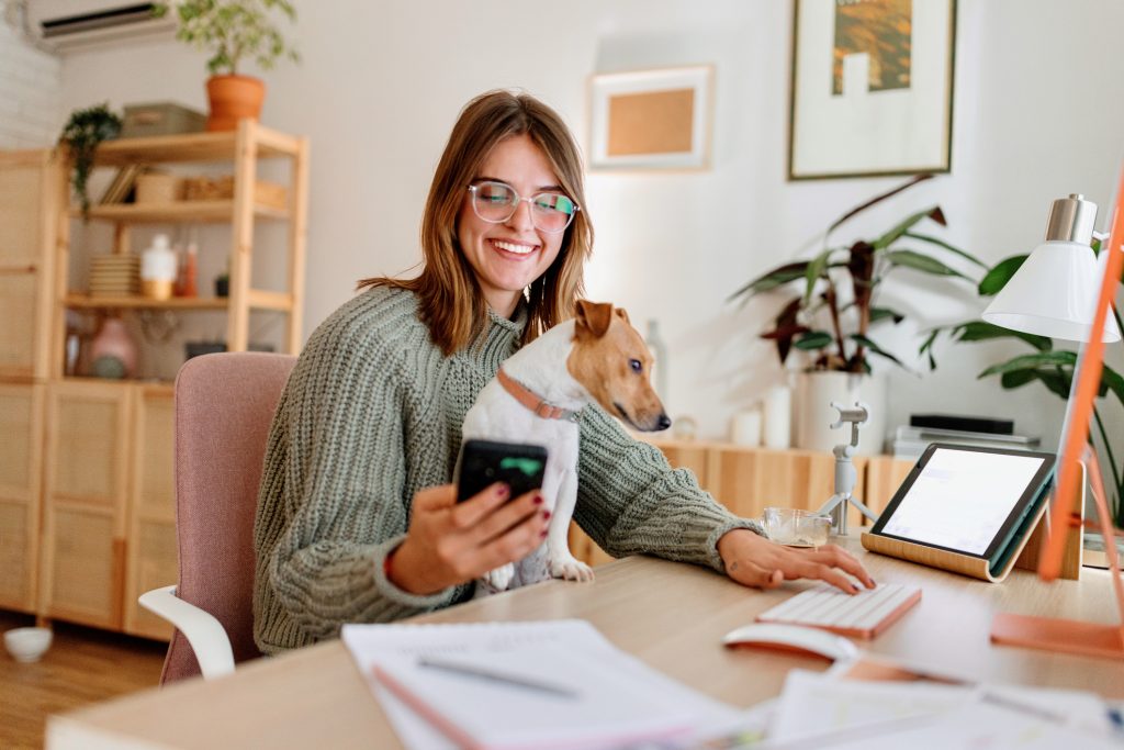 A woman, wearing glasses and a sweater, looks at her phone while a small dog sits on her lap at a wooden desk. The desk, cluttered with papers, a tablet, and office supplies, hints she might be researching local dog daycare options.