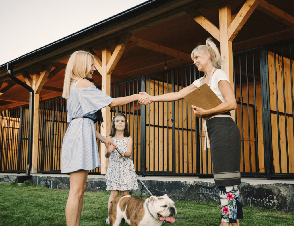 Two women shake hands outside a wooden building as a child claps nearby. A bulldog, perhaps booked in using the latest all-in-one dog boarding software, sits contentedly on the grass while one woman holds a clipboard.