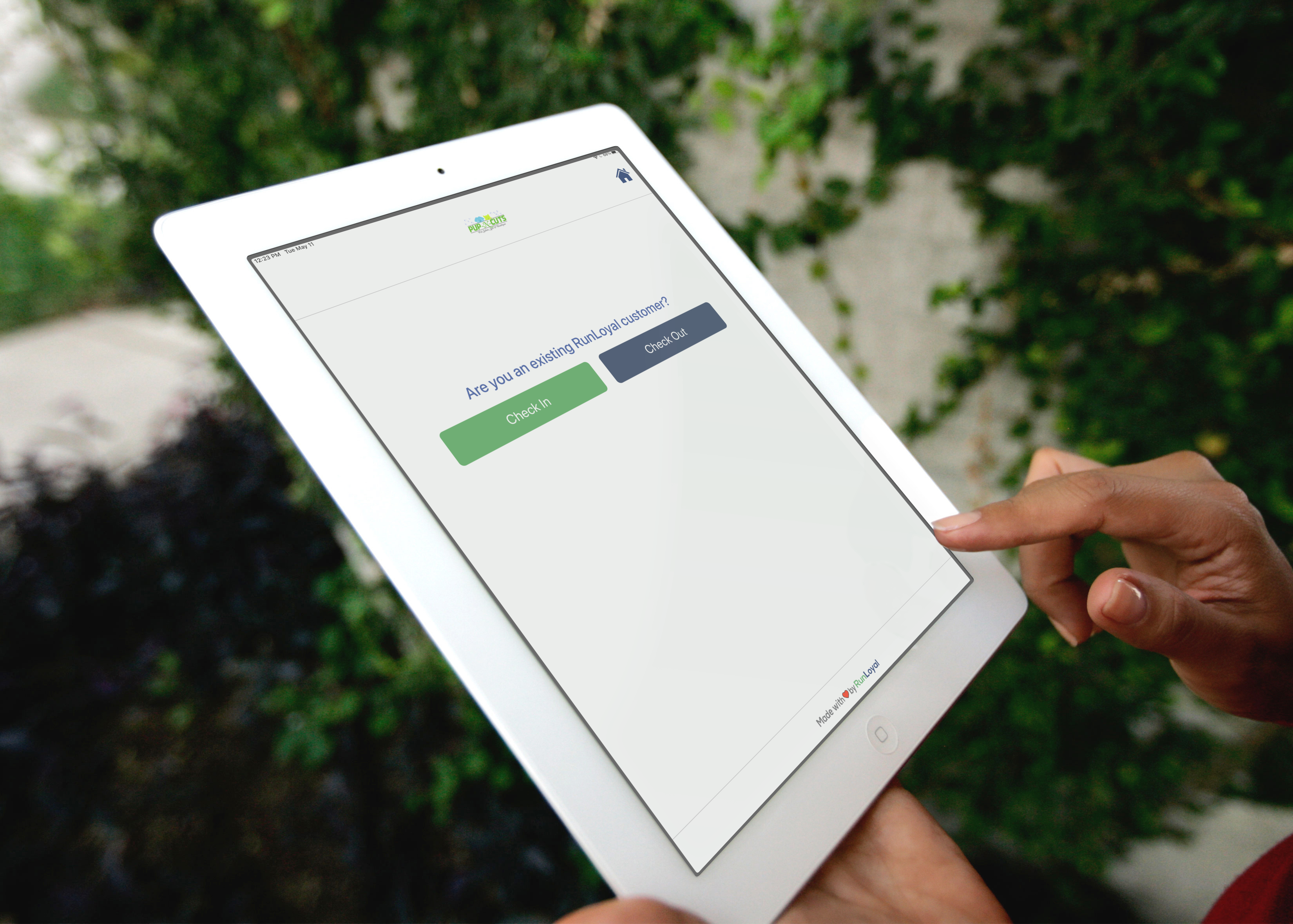 A person uses a tablet displaying a question about clocking in or out, with buttons for Clock In and Clock Out. The background of lush greenery sets the scene, perfect for businesses like RunLoyal | Dog Business Software | Boarding, Daycare to manage their operations smoothly.